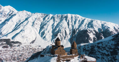 Gergeti trinity church sits on a hilltop overlooking the town of stepantsminda and the towering, snow-capped caucasus mountains in the distance, creating a breathtaking winter scene in georgia