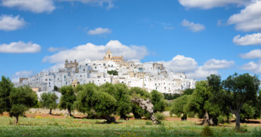 Landscape in Puglia, Italy, with the white city (citta bianca) Ostuni on a hill above an olive tree orchard under a cloudy sky.