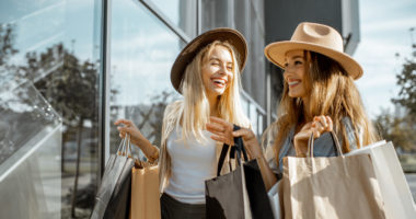 Two happy girlfriends looking on the shopwindow while standing with shopping bags near the mall