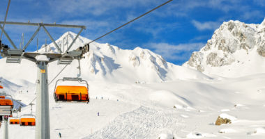 Ski lift ropeway on hilghland alpine mountain winter resort on bright sunny day. Ski chairlift cable way with people enjoy skiing and snowboarding.Banner panoramic wide view of downhill slopes.